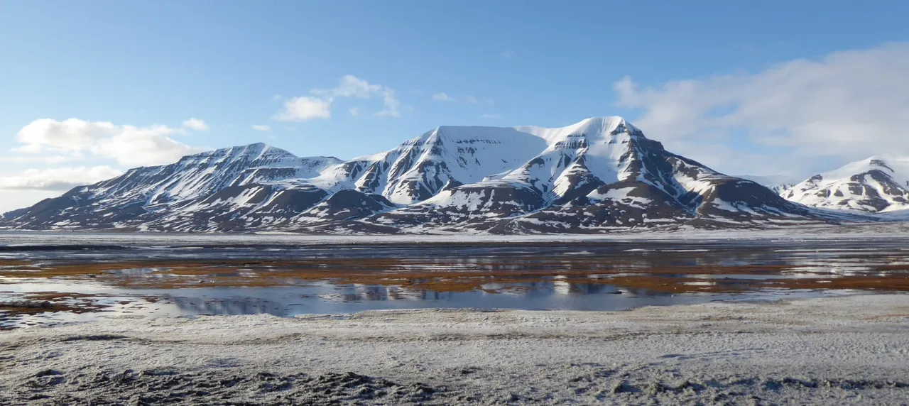 Snow-streaked mountains rising behind a wide, partly frozen tidal flat under a blue sky