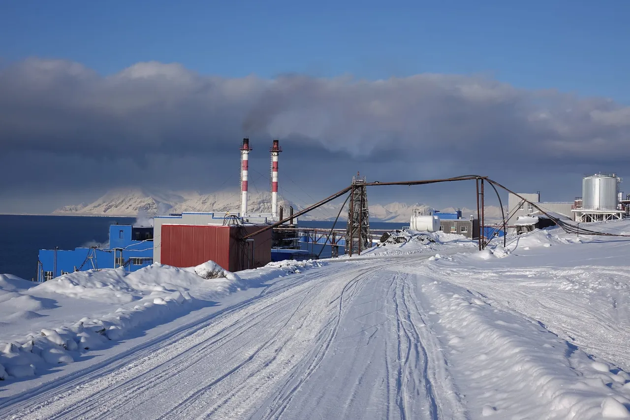 A snow-covered road leading past the red and blue buildings of Barentsburg's coal power station, with two tall striped chimneys and a steaming stack