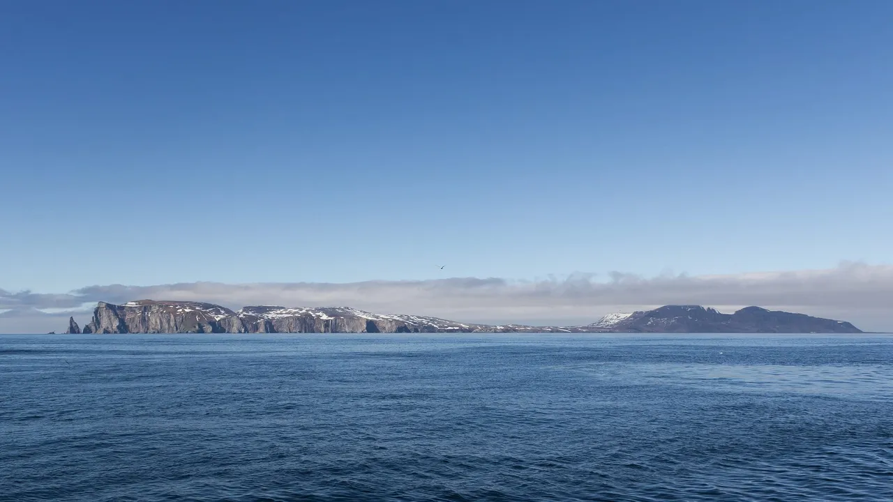 A long, flat-topped island with steep cliffs at each end, seen across calm blue sea under a clear sky