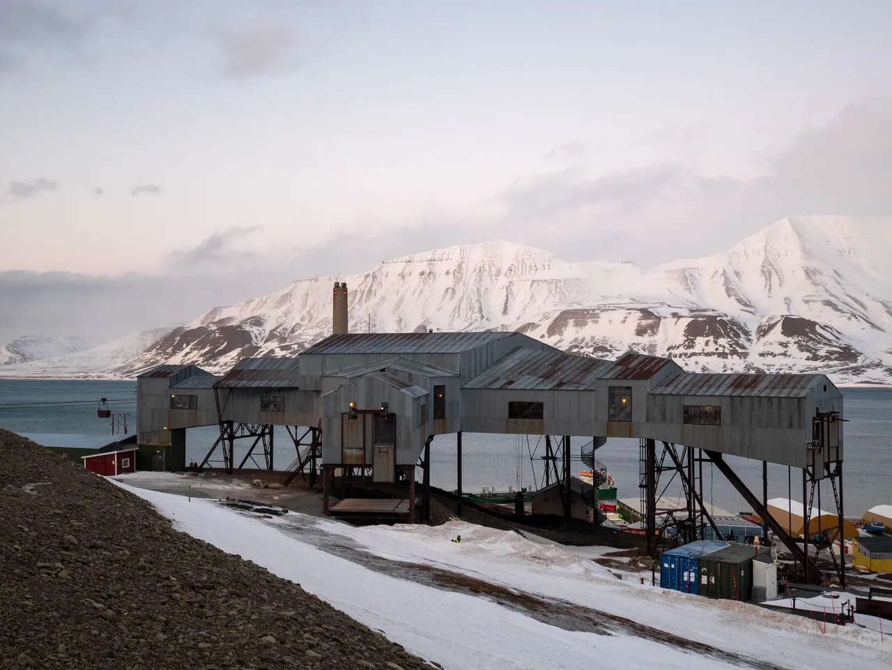 The grey timber buildings of the old coal cableway centre standing on stilts above the town, with snowy mountains across the fjord behind