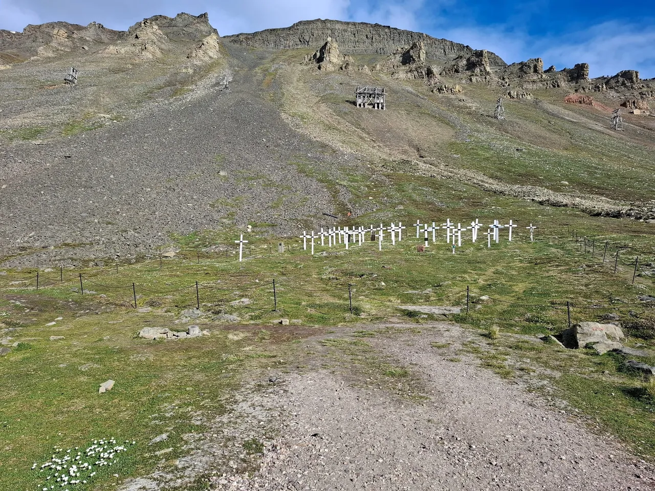 Rows of white wooden crosses inside a low fence on a green slope, with a grey mountainside and an old mine structure above