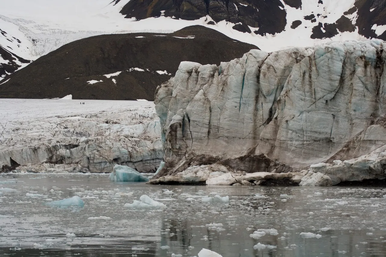 The high, cracked front of a glacier meeting calm grey water dotted with floating ice, below dark mountains