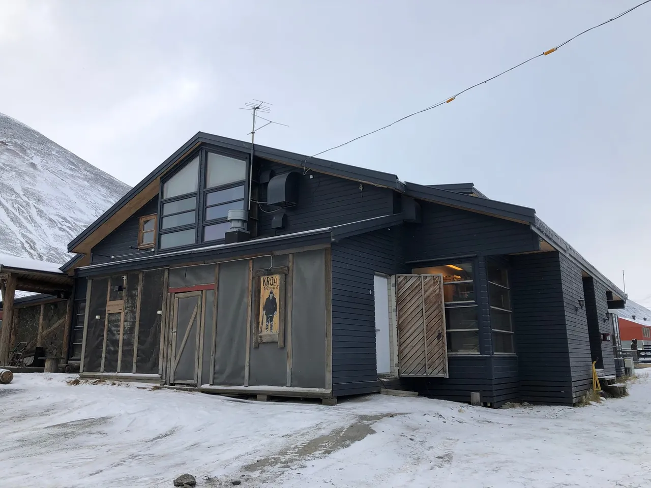The dark timber front of Kroa, with large windows and a covered entrance, on a snowy street below a mountain