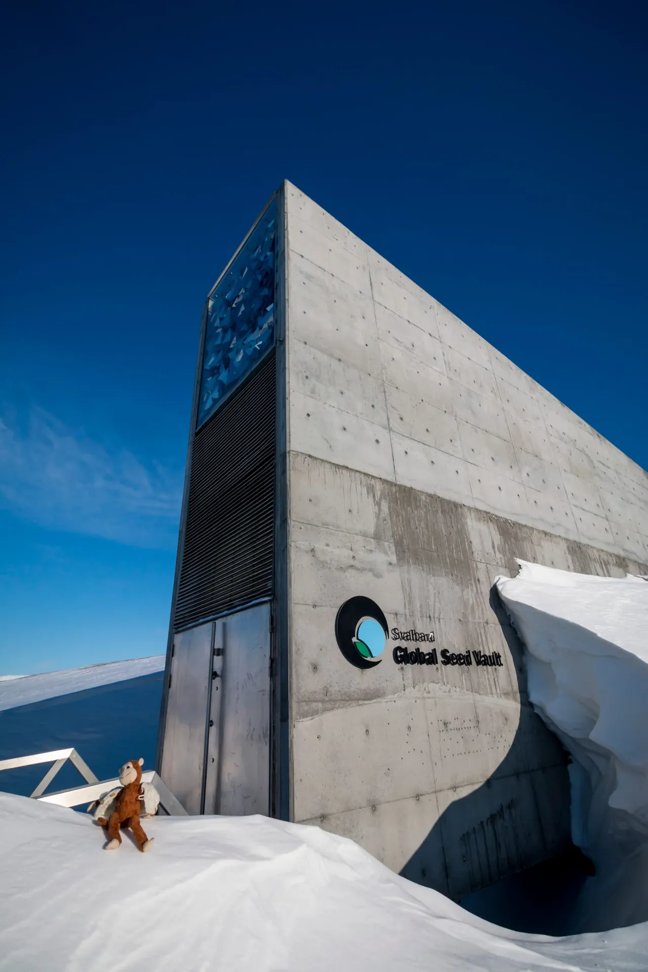 The angular concrete entrance of the Seed Vault rising out of deep snow under a blue sky, with a panel of reflective artwork at the top