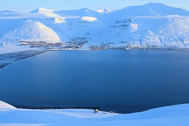 Longyearbyen's houses strung along the far shore of a dark blue fjord, beneath snow-covered mountains in low spring light