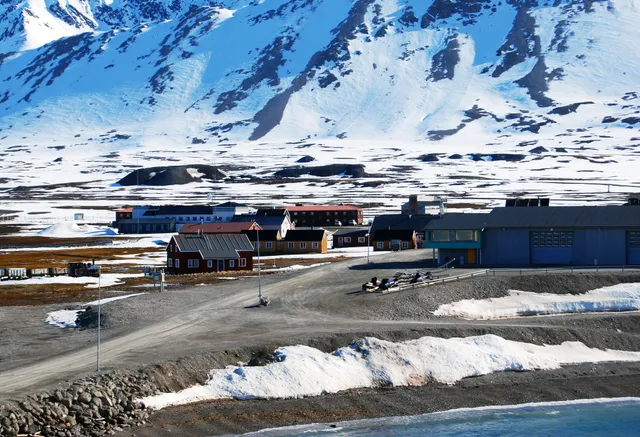 Brightly painted wooden houses and a blue warehouse by the shore at Ny-Ålesund, below a steep snow-covered mountainside