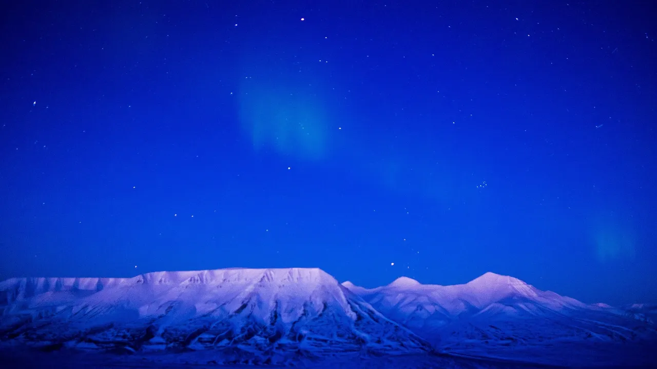 Snow-covered mountains glowing pale pink and violet under a deep blue sky with a faint aurora and stars