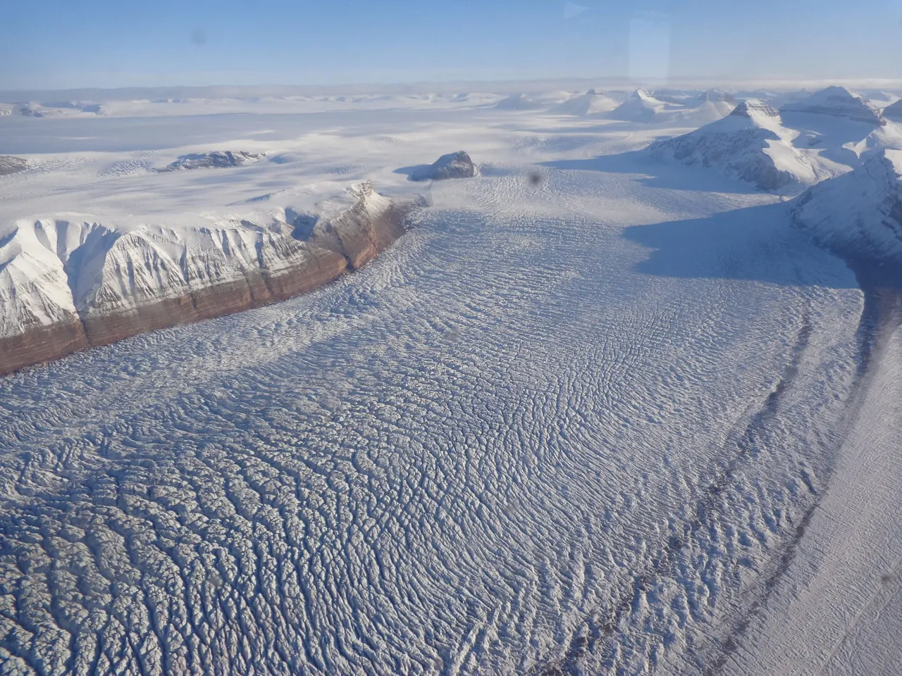 A vast glacier seen from above, its surface broken into a maze of crevasses, flowing between dark mountain ridges