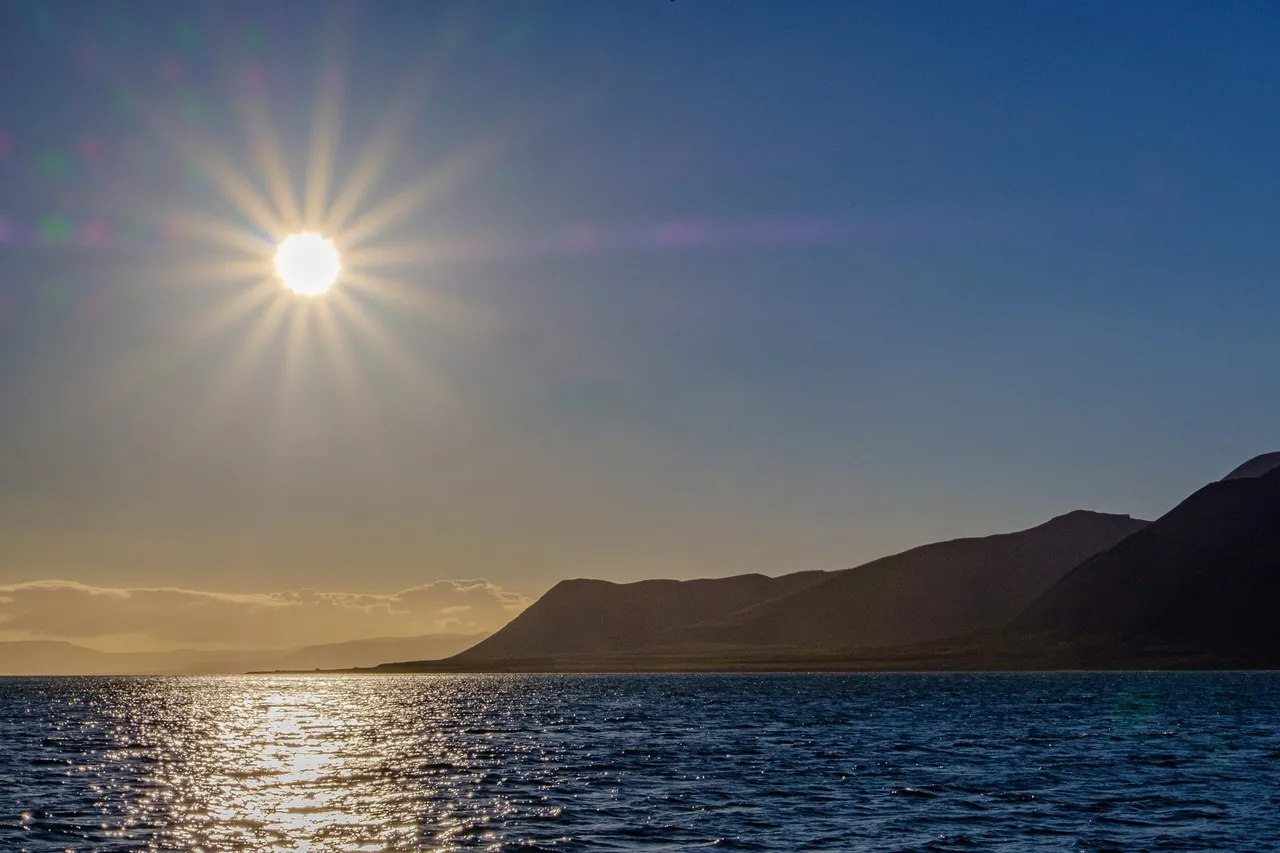 A bright sun low over a dark fjord, its light glittering across the water below a mountain ridge