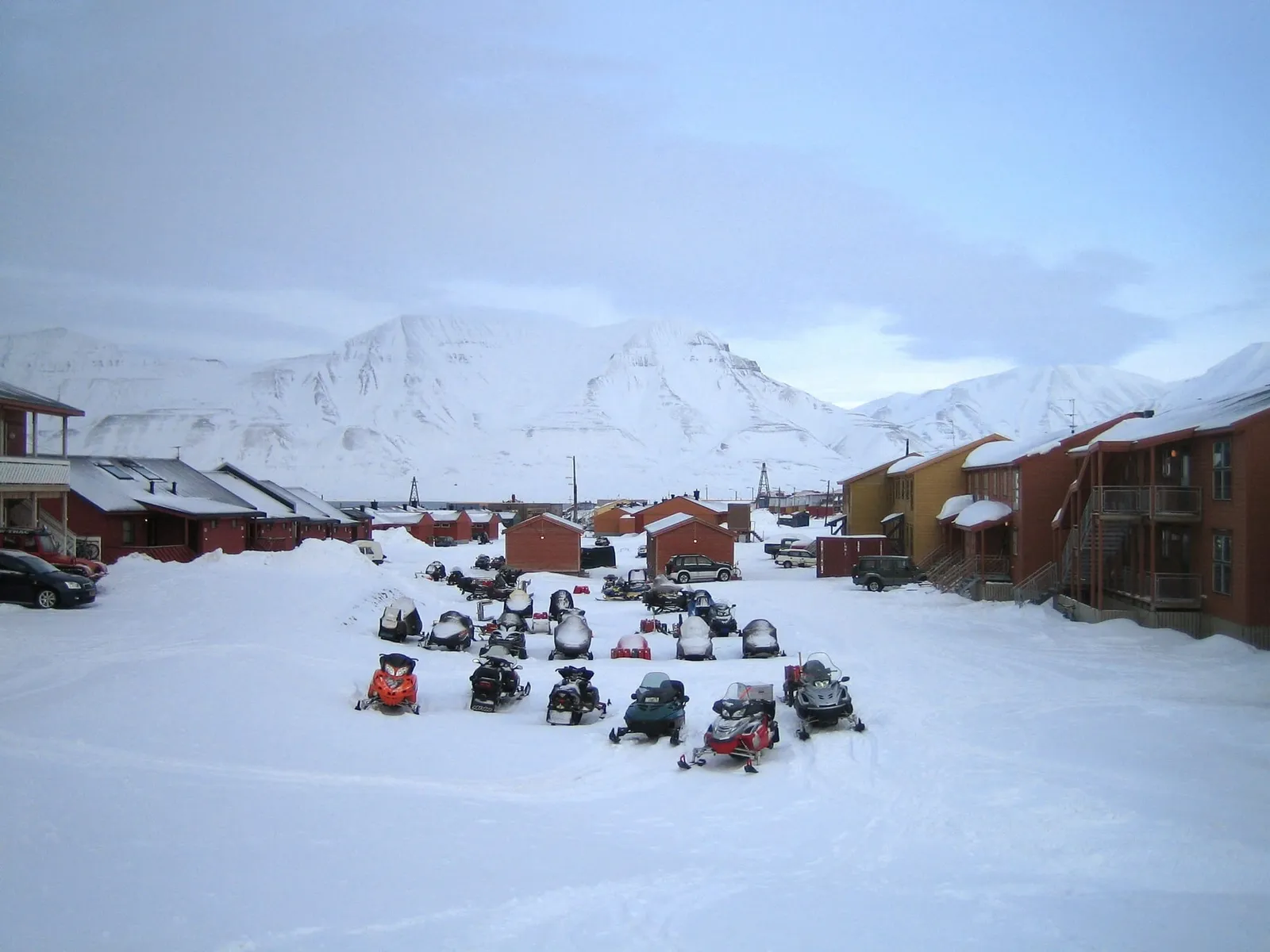 Dozens of snowmobiles parked in rows on the snow between wooden houses in Longyearbyen, with white mountains behind
