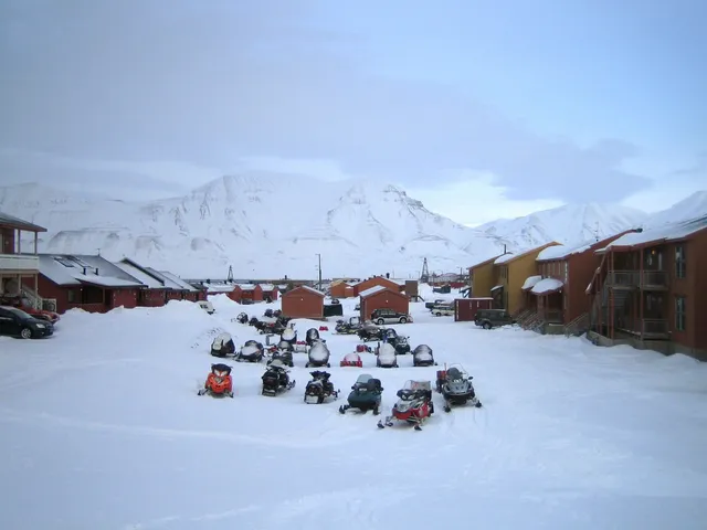 Dozens of snowmobiles parked in rows on the snow between wooden houses in Longyearbyen, with white mountains behind