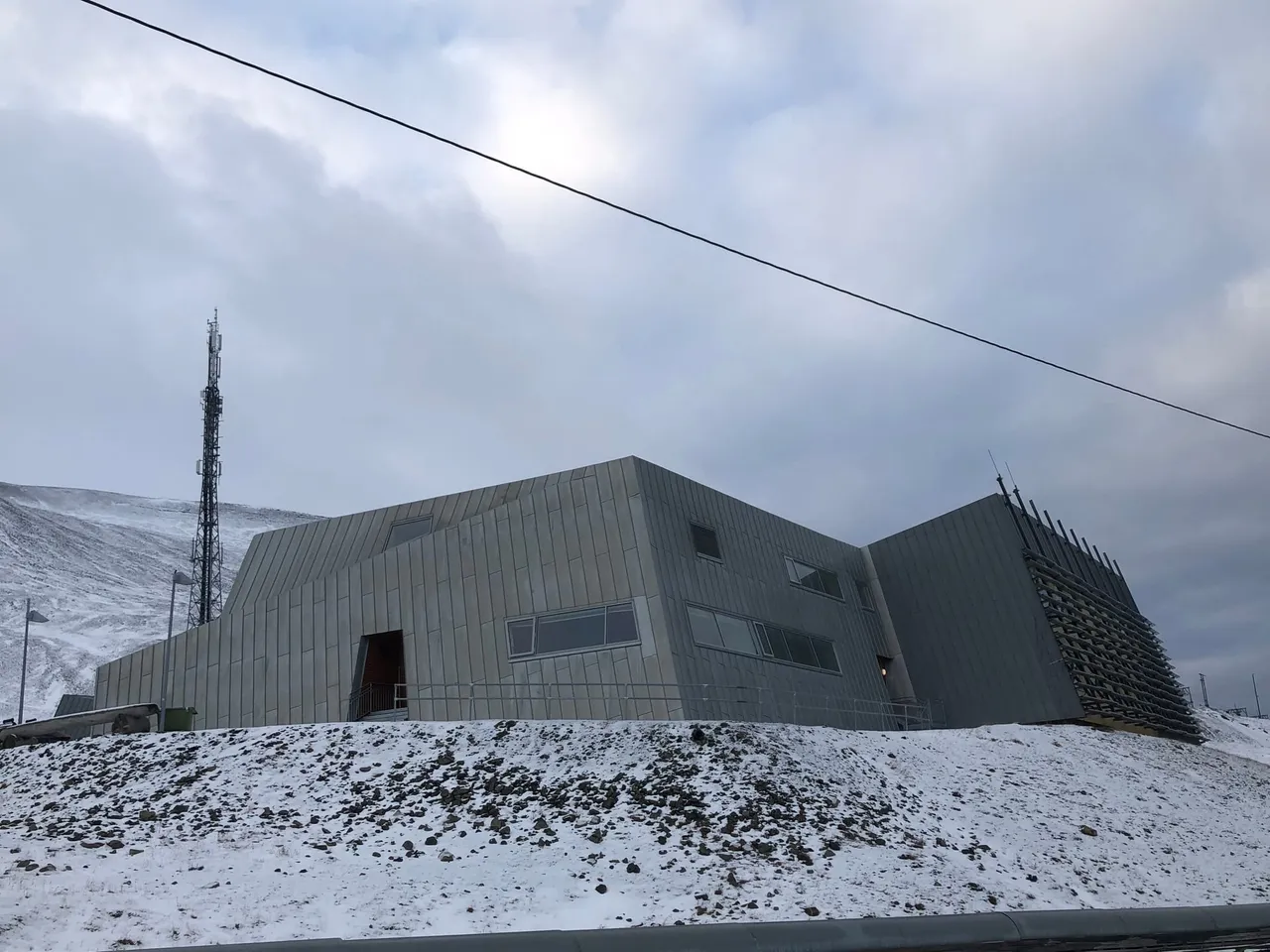 A modern grey building with angular walls and a tall radio mast, on a snowy slope under a cloudy sky